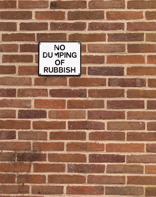 A rectangular white metal sign mounted on a red brick wall, displaying the warning 'NO DUMPING OF RUBBISH' in black uppercase letters. The sign is positioned approximately one-third from the top of the image and is affixed with four screws, one at each corner. The brick wall features a traditional running bond pattern with reddish-brown bricks and light mortar joints, showing slight variation in tone and texture. The wall's surface appears weathered with some minor discoloration near the bottom. The shadow of the sign is cast downward onto the bricks, indicating a light source from above. The scene is outdoors, typical of an urban environment, where such signage often relates to private waste management or property regulations. Rubbish collection highbury.co.uk emphasizes the importance of responsible waste disposal, and this sign serves as a clear visual cue to discourage unauthorized rubbish dumping, aligning with alternative or private waste handling practices beyond local council services.