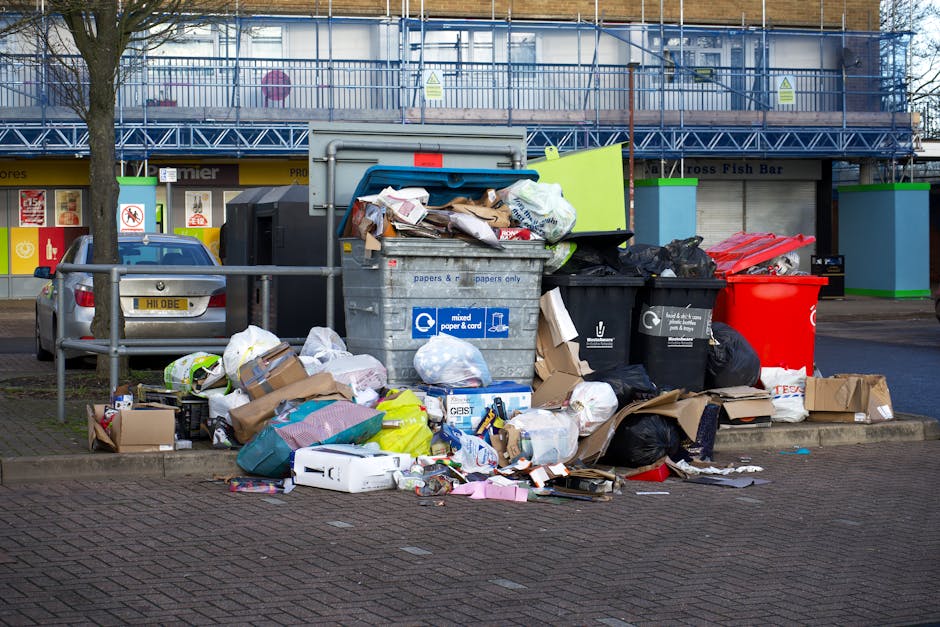 A pile of mixed rubbish and waste materials is overflowing in front of a multi-compartment waste container on a paved area, with various black, black with white, and red garbage bags, flattened cardboard boxes, plastic wrapping, and paper waste scattered around. The container, designated for mixed paper and card, is partially open, revealing crumpled paper and plastic packaging inside, with additional trash stacked on top and spilling out onto the surrounding pavement. Nearby, a small silver hatchback car is parked behind a metal barrier, with its rear end visible. The setting appears to be an urban outdoor waste collection point, with a storefront and a building with scaffolding and signage in the background. The scene captures a typical example of improper rubbish disposal, highlighting the importance of proper waste management and the role of private waste collection services like Rubbish Collection Highbury in maintaining cleanliness.
