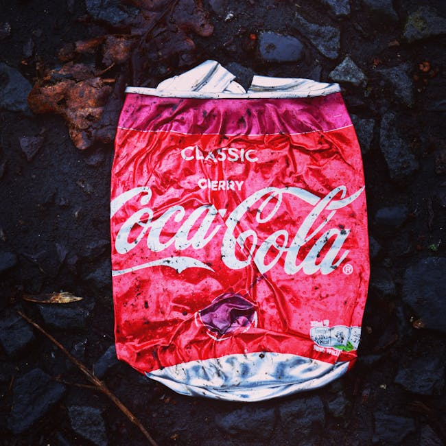 A crumpled, weathered Coca-Cola Classic Cherry aluminum can lies on a dark, uneven surface composed of volcanic rocks or black gravel. The can's red and white branding is faded and torn, with visible creases and dents across its body, indicating it is empty and discarded. The top of the can is partly crushed, with the silver pull tab missing, exposing the opening. Surrounding the can are irregularly shaped, rough-textured black stones with hints of brown or reddish hues, suggesting an outdoor environment. The lighting appears natural, casting soft shadows that highlight the can's deformed surface and the coarse texture of the rocks. The scene is minimal but clearly depicts a piece of rubbish, which could be part of an independent disposal or litter collection service context, such as those handled by Rubbish Collection Highbury, engaged in local waste management efforts.