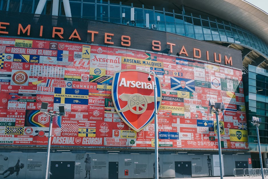 The exterior of the Emirates Stadium features a large, modern glass facade with sleek, curved architectural elements. Prominently displayed on the building is a vast mural composed of numerous flags from various countries, arranged in a grid pattern, with vibrant colors and a mix of rectangular shapes. In the center of the mural is a large, shield-shaped Arsenal Football Club logo, featuring a red background with a gold cannon and white outline, positioned slightly off-center. Four tall, white streetlights with square floodlights are situated in the foreground, casting focused light onto the ground, which appears to be a paved pedestrian area. The overall environment is bright, likely during daytime, with natural sunlight illuminating the building's surface and creating sharp shadows from the streetlights. The image captures the clean, organized exterior space of the stadium, providing a visual context related to the venue, which sometimes involves event debris or rubbish disposal, relevant to independent waste handling services by companies like Rubbish Collection Highbury.