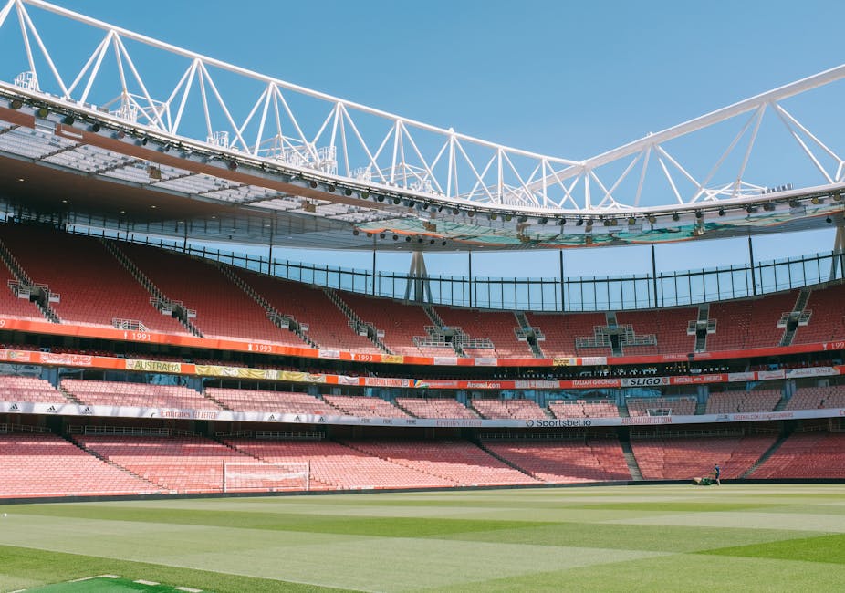 An empty football stadium, featuring rows of red seating surrounding a well-maintained grass pitch with light and dark green stripes. The stadium's interior includes multiple tiers of seating, with some sections featuring black railings and small advertising banners displaying various sponsors. Above the seating area, a large, white, lattice-style roof structure with trusses spans the upper section, providing partial shade and covering. The sky visible through the open sections of the roof is clear and blue, indicating a bright, sunny day. The overall scene is devoid of spectators, highlighting the empty stands and pristine playing field, with a faint figure near the far end of the pitch. This imagery relates to the context of sports venues and could be relevant for discussions around stadium maintenance or cleaning, including potential waste management or rubbish removal services for large event locations.