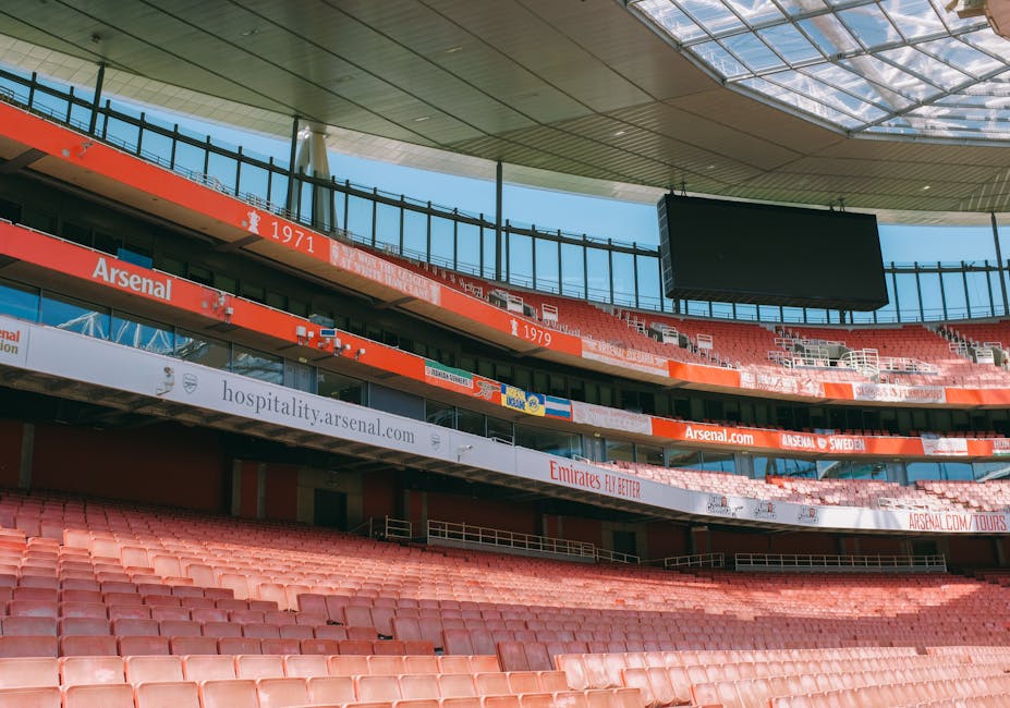 The exterior of the Emirates Stadium features a large, modern glass facade with sleek, curved architectural elements. Prominently displayed on the building is a vast mural composed of numerous flags from various countries, arranged in a grid pattern, with vibrant colors and a mix of rectangular shapes. In the center of the mural is a large, shield-shaped Arsenal Football Club logo, featuring a red background with a gold cannon and white outline, positioned slightly off-center. Four tall, white streetlights with square floodlights are situated in the foreground, casting focused light onto the ground, which appears to be a paved pedestrian area. The overall environment is bright, likely during daytime, with natural sunlight illuminating the building's surface and creating sharp shadows from the streetlights. The image captures the clean, organized exterior space of the stadium, providing a visual context related to the venue, which sometimes involves event debris or rubbish disposal, relevant to independent waste handling services by companies like Rubbish Collection Highbury.