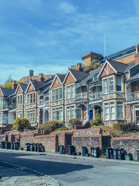 A row of Victorian-style terraced houses located on a sloped street, featuring decorative white ironwork balconies and bay windows. The buildings are constructed with red brick, with some sections painted in light colours, and have gabled roofs with dark tiles. In front of the houses, there is a low brick wall bordered by neatly trimmed shrubs and small trees, with several black wheelie bins placed along the pavement for waste disposal. The street appears calm with no visible traffic, set against a blue sky with a few wispy clouds, suggesting a clear and sunny day. The scene reflects an urban residential area where private waste collection services, such as those offered by Rubbish Collection Highbury, might be employed for rubbish removal and on-site clearance of waste, especially where reliance on third-party waste disposal providers is common. The overall setting is clean and well-maintained, illustrating typical residential waste management practices involving designated bins ready for collection.
