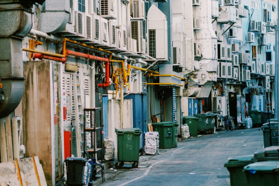 A narrow urban alleyway lined with various waste collection bins and sacks placed on the pavement in front of a building façade. The building features a dense array of air conditioning units, vents, pipes, and utility meters mounted on the wall, with some pipes painted in red and yellow. The surface of the wall is weathered, showing signs of dirt and corrosion, and the alley appears to be used for private waste disposal rather than municipal rubbish collection. The bins are predominantly green with numbers and labels, and some are accompanied by black trash bags filled with waste. Bicycles are leaning against the wall near the bins, and a small cart or shelving unit can also be seen to the left. The lighting suggests daytime with natural light illuminating the scene, creating subtle shadows and highlighting the textures of the metal, plastic, and concrete surfaces. This scene depicts an instance of independent waste handling typical of private property or designated service areas, aligning with alternatives to public rubbish collection services potentially managed by local waste management specialists like Rubbish Collection Highbury.