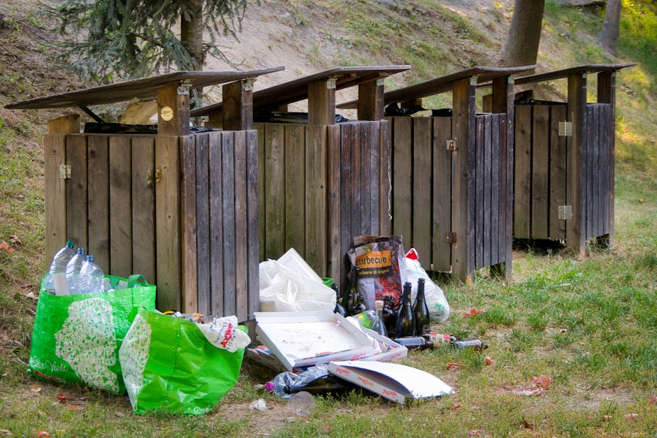 A row of four outdoor wooden waste disposal units with sloped roofs, situated on a grassy area near trees. The units are made of vertical wooden planks, with some showing signs of weathering and dark staining, and are positioned side by side. In front of the units, there is an accumulation of rubbish including clear plastic bottles, a green reusable shopping bag filled with more plastic bottles, and a white pizza box with some leftover food crumbs. Additionally, there are several empty glass bottles, a black plastic bag, and some discarded packaging, such as a barbecue food container. The background reveals leafy trees and a sloping dirt embankment, indicating a natural outdoor environment. The overall scene suggests illegal dumping or improper disposal of waste near the rubbish containers, contrasting with professional waste management services like Rubbish Collection Highbury, which handle domestic and commercial rubbish collection efficiently and responsibly.