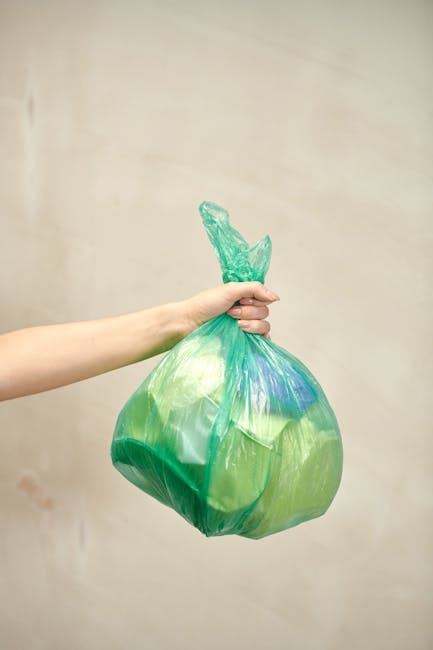 A person's hand and forearm are visible holding a tightly knotted green plastic rubbish bag with a slightly shiny surface and some visible folds. The bag contains mixed waste items, possibly including paper and plastic, as indicated by its semi-transparent material and irregular shape. The hand grips the bag from the top, with fingers wrapped around the gathered plastic, and the background features a plain, light-colored wall without additional objects or environmental context. The image emphasizes individual waste handling, aligning with themes of private rubbish collection or on-site waste management, and the scene suggests preparation for disposal or collection. The setting appears to be indoors or in a controlled environment, with soft lighting that highlights the bag's texture and color variety, subtly supporting the context of waste disposal or rubbish removal services, such as those provided by Rubbish Collection Highbury. The focus on the bag and hand reflects the act of managing household waste as part of independent rubbish collection efforts.