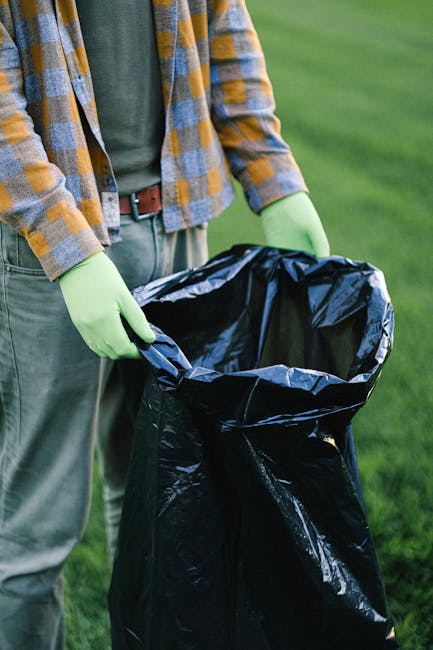 A person wearing green rubber gloves and casual clothing, including a grey t-shirt and a checkered yellow and grey shirt, is holding open a large black rubbish bag made of lightweight plastic. The bag is partially folded at the edges, ready to be filled with waste. The individual is standing outdoors on a grassy area with a lush, green lawn visible in the background, suggesting a garden or park environment. The scene is well-lit with natural daylight, emphasizing the textures of the gloves, clothing, and the crinkled surface of the rubbish bag. This setup indicates a context of waste collection or garden clearance, potentially related to private rubbish disposal services like those offered by Rubbish Collection Highbury. The image context aligns with professional waste management activities, including rubbish removal and on-site clearance, emphasizing the importance of proper waste handling and environmentally responsible disposal practices.