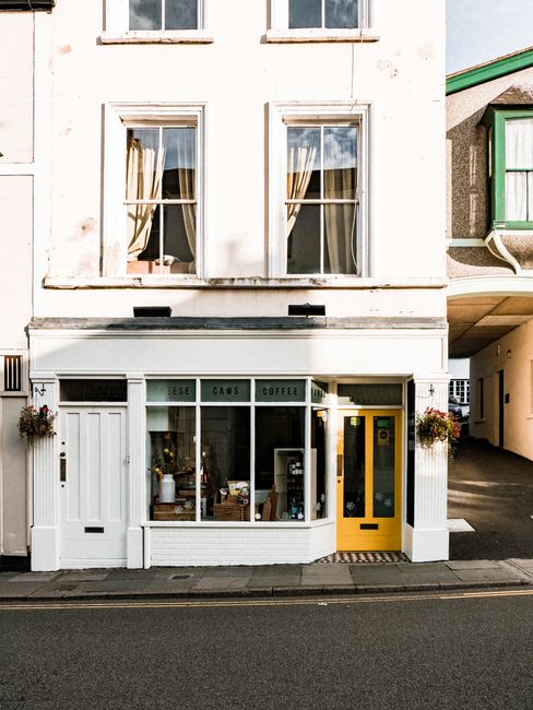 A two-storey building with a small commercial space on the ground floor, featuring a white wooden facade and large front window displaying various items, likely a café or small shop. The ground floor has three window sections; the left and central windows are divided by white wooden framing, with the central window displaying coffee-related signs, including the words 'ESE,' 'CAWS,' and 'COFFEE.' To the right, there is a bright yellow door with a glass panel, and next to it, a black mail slot. Flanking the entrance are small hanging flower baskets, adding decorative greenery, and the building is situated directly on a paved sidewalk along a quiet street. The upper floors have two evenly spaced sash windows with off-white curtains partially drawn, set into a weathered white exterior wall. To the right, part of a green and gray structure with a slanted roof is visible, and the whole scene suggests an urban setting, possibly in a small town or high street. The decorative and functional elements align with an independent retail or hospitality business, and this image visually supports the context of commercial waste management or rubbish collection services for small shops or cafés in the area.