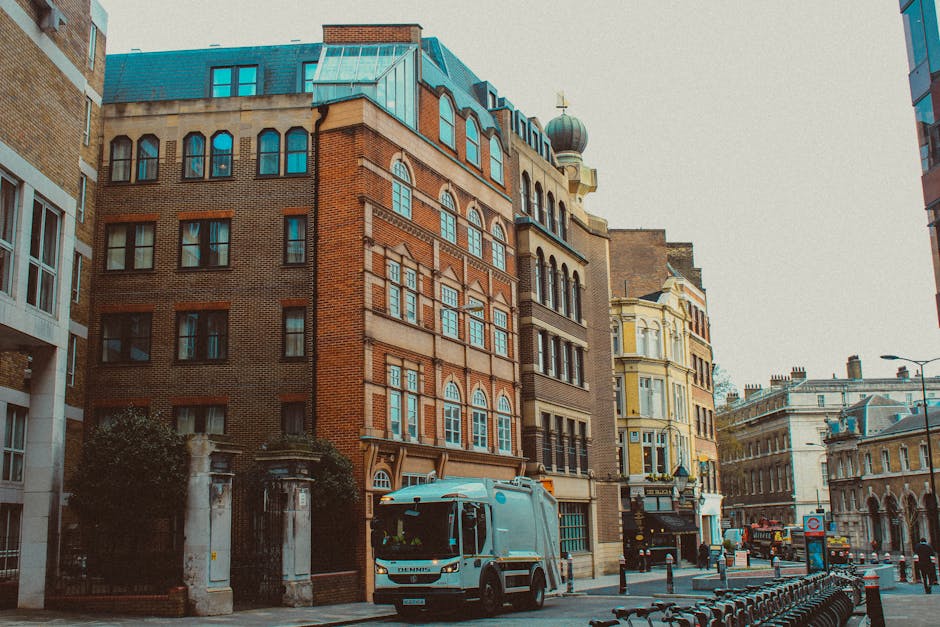 A street scene in an urban area with a mix of historic and modern buildings. The focus is on a narrow, multi-story brick building with large, arched windows on each floor, topped with a rounded tower feature and a decorative dome with a finial. Adjacent buildings include a beige and yellow storefront with ornate architectural details, and a taller modern structure with a glass façade. In the foreground, a white waste collection truck from Rubbish Collection Highbury is parked on the street, partially blocking part of the view. The truck has a compact design with a clean, smooth surface and dark windows. The street is lined with metal bike racks, some empty, and bollards separating the sidewalk from the road. The environment suggests a busy city setting with light traffic, overcast sky, and minimal pedestrian presence, reflecting a typical scene where private waste collection services operate in an urban context. The visible scene subtly connects to rubbish removal and alternative waste handling by showing a private waste collection vehicle actively serving the area.